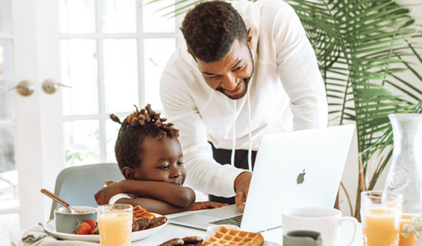 Father and son sit at the breakfast table looking at a computer together.