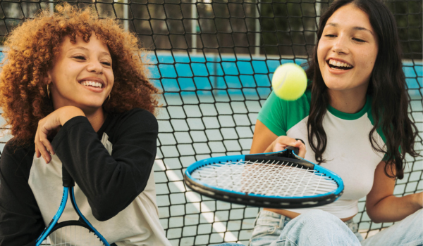 Two teenage girls sit on a tennis court after a parks and rec program. 