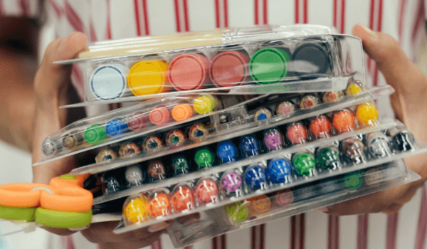 A staff member holds a stack of art supplies (pens, markers, paint, etc.)