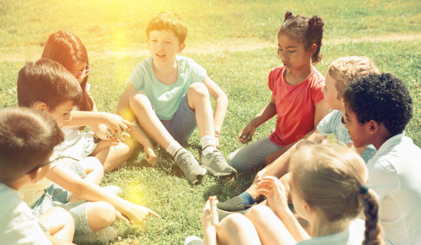 A group of children sit in a circle outside at a YMCA summer camp. 